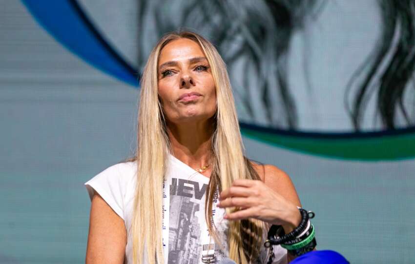 Adriane Galisteu sentada no palco, de camiseta branca, mexendo nos cabelos com a mão esquerda