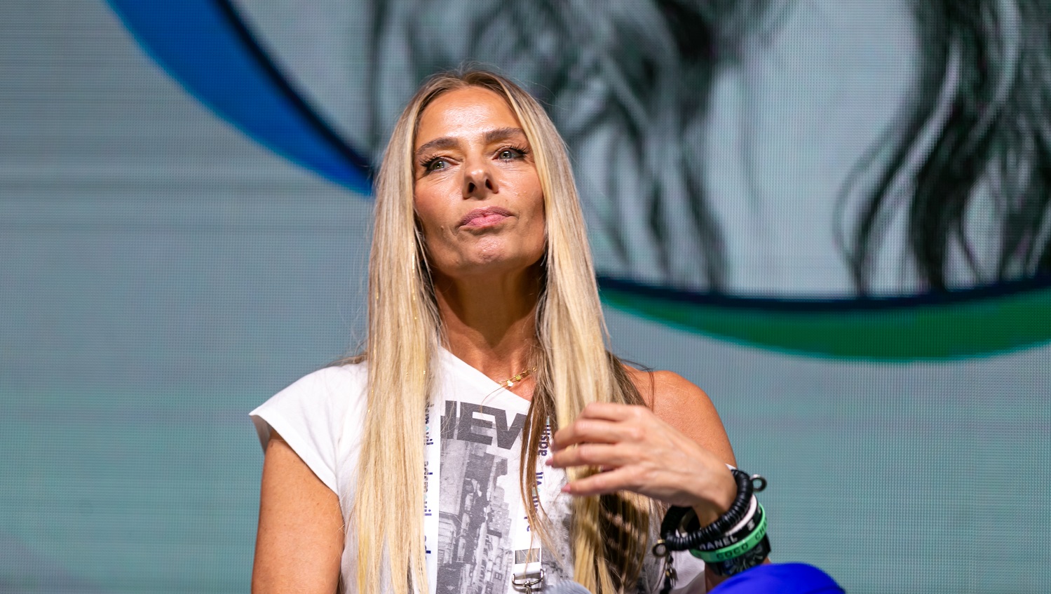 Adriane Galisteu sentada no palco, de camiseta branca, mexendo nos cabelos com a mão esquerda