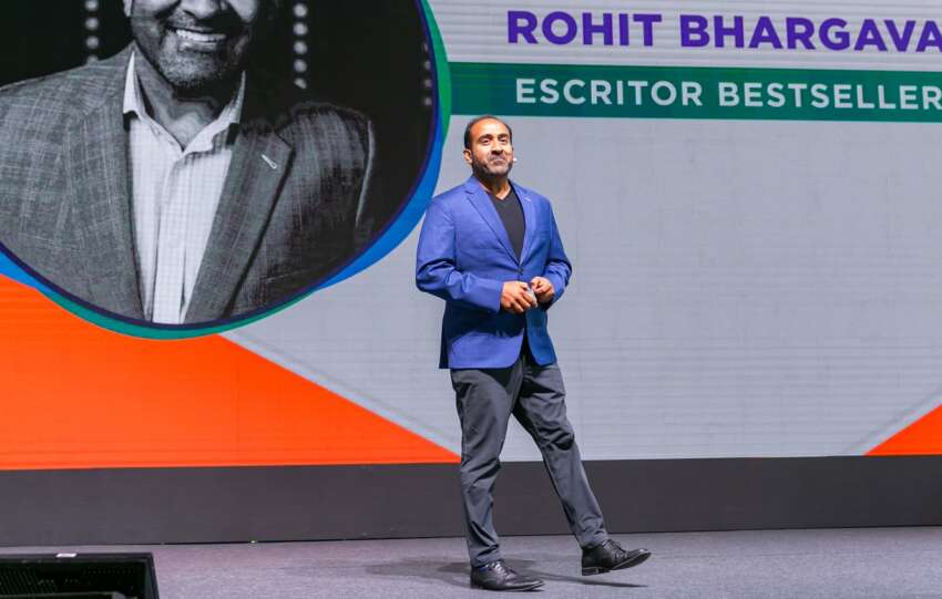 Rohit Bhargava em cima do palco de blazer azul e calça cinza, em frente ao telão com seu nome e foto