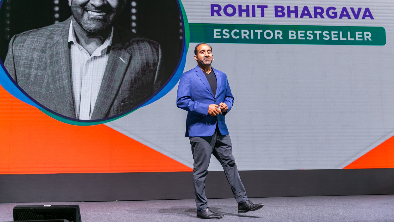 Rohit Bhargava em cima do palco de blazer azul e calça cinza, em frente ao telão com seu nome e foto