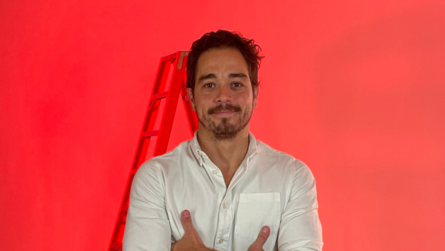 Guilherme Barsanti Leitede camisa branca e braços cruzados, à frente de uma parede vermelha com uma escada